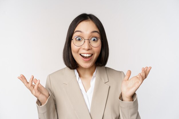 Enthusiastic asian saleswoman in glasses smiling and laughing looking amazed at camera standing in beige suit over white background