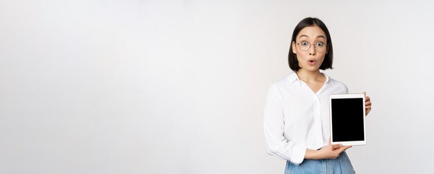 Enthusiastic asian woman office worker in glasses showing digital tablet screen demonstrating info on gadget display standing over white background