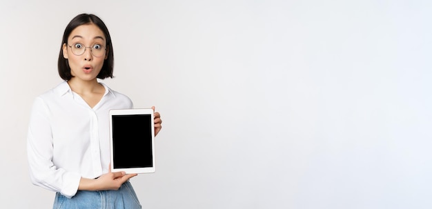 Восторженная азиатская офисная работница в очках, показывающая экран цифрового планшета, демонстрирующая информацию о дисплее гаджета, стоящем на белом фоне