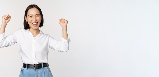 Enthusiastic asian woman rejoicing say yes looking happy and celebrating victory champion dance fist pump gesture standing over white background