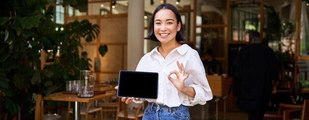 無料写真 熱心なアジアの女性は、タブレットの画面に大丈夫のサインを示し、感心しているように見えます