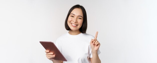 Enthusiastic asian woman with tablet raising finger and looking amazed pointing up standing over whi