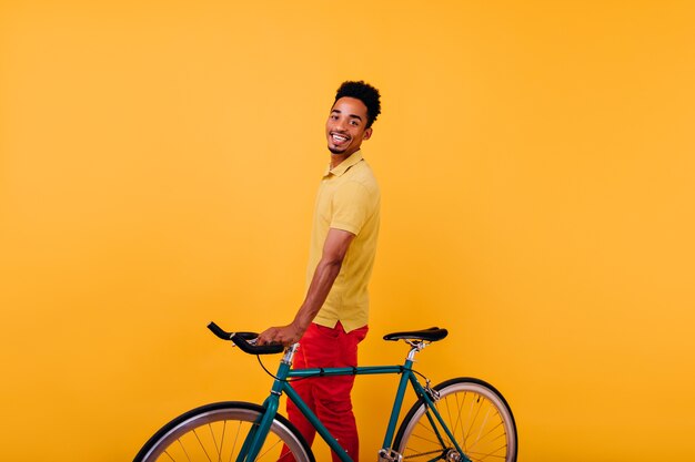 自転車でポーズをとる陽気な笑顔で熱狂的な黒人男性。自転車で立っている楽しいアフリカの男性モデルの屋内写真。