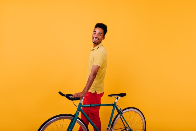 자전거와 함께 포즈를 취하는 밝은 미소로 열정적 인 흑인 남자. 자전거와 함께 서있는 즐거운 아프리카 남성 모델의 실내 사진.