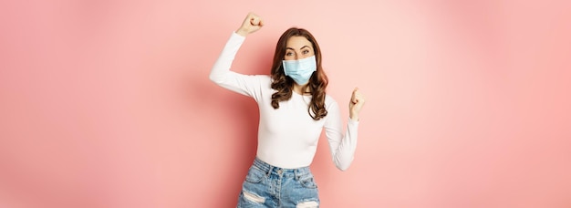 Free photo enthusiastic brunette girl in medical face mask dancing and laughing celebrating victory triumphing
