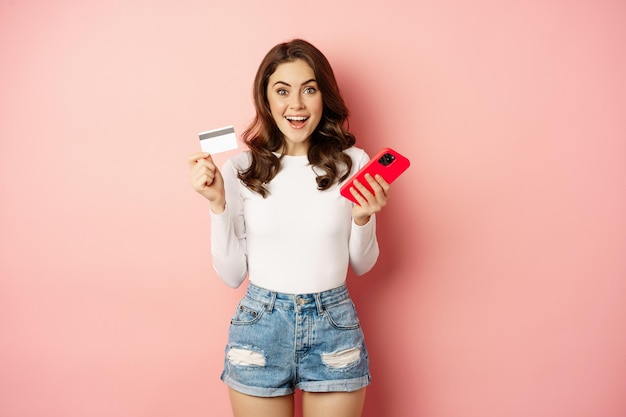 Восторженная брюнетка показывает кредитную карту и использует мобильный телефон для заказа или оплаты, приложение для покупок в Интернете, стоя на розовом фоне