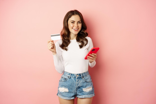 Enthusiastic brunette girl showing credit card and using mobile phone to order or pay, online shopping app, standing over pink background