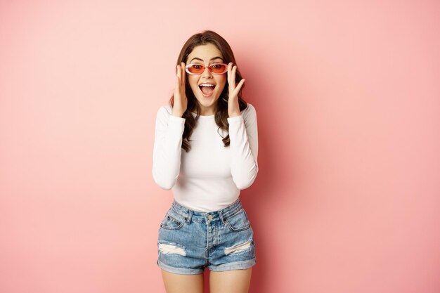 Enthusiastic brunette girl in stylish sunglasses reacts surprised and amazed looking with disbelief ...