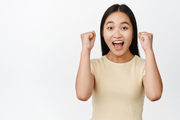 Enthusiastic brunette girl yelling yes clench fists and celebrating winning something standing in yellow tshirt over white background