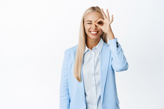 Enthusiastic businesswoman showing zero okay gesture on eye and smiling recommending something good standing in suit over white background