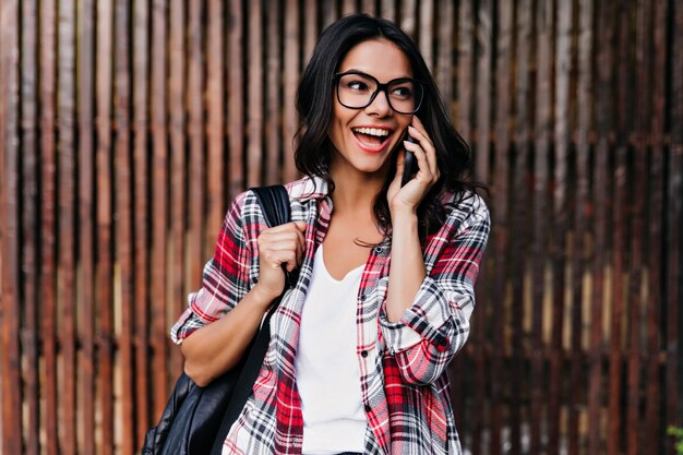 Enthusiastic caucasian girl in glasses talking on phone with smile. Outdoor portrait of blithesome brunette woman posing with leather backpack.