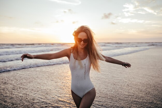 熱狂的な白人の女の子が海の海岸を駆け下りて笑っています。エキゾチックなリゾートで夏休みを楽しんでいる白い水着の嬉しい女性の屋外の肖像画。