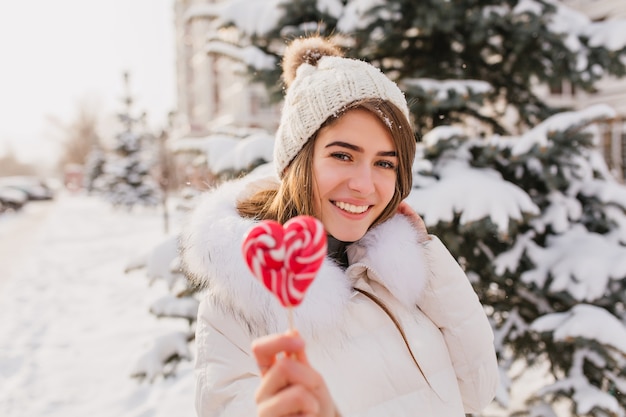 Восторженная кавказская женщина, держащая леденец на палочке сердца во время зимней фотосессии. Счастливая женщина в вязаной шапке и белом халате позирует со сладкими конфетами во время работы в снежном парке.