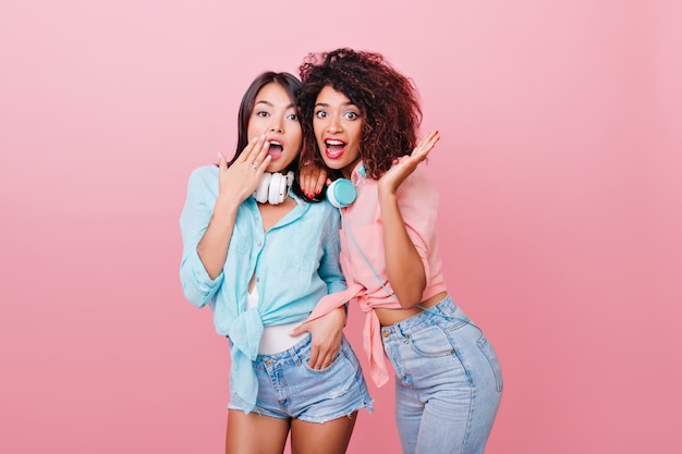 Enthusiastic caucasian woman with surprised face expression posing near mulatto curly girl. Indoor photo of shocked ladies in colorful shirts standing.