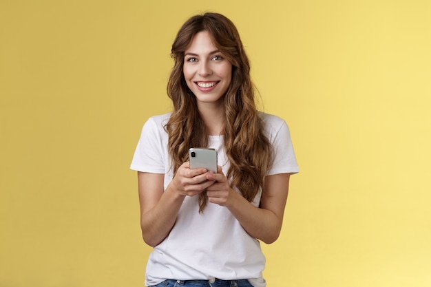 Enthusiastic charming sociable young girl messaging friend sending photos social media hold smartphone look camera happily friendly smiling stand yellow background casual outfit