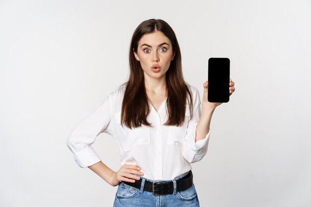 Enthusiastic corporate woman demonstrating website, mobile phone screen, showing application and saying wow, standing over white background.