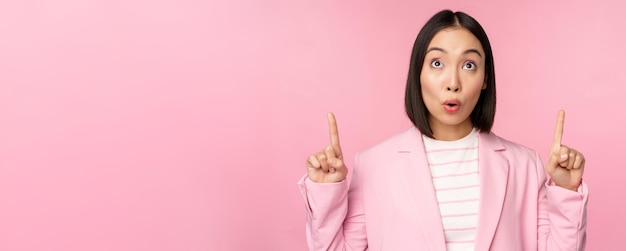 무료 사진 열정적인 기업 직원 아시아 비즈니스 여성이 손가락을 가리키고 분홍색 배경 위에 서 있는 광고 로고를 보여주며 웃고 있습니다.