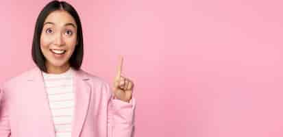 無料写真 ピンクの背景の上に立っている広告のロゴを表示して指を上に向けて笑顔で熱狂的な企業の労働者アジアのビジネスウーマン