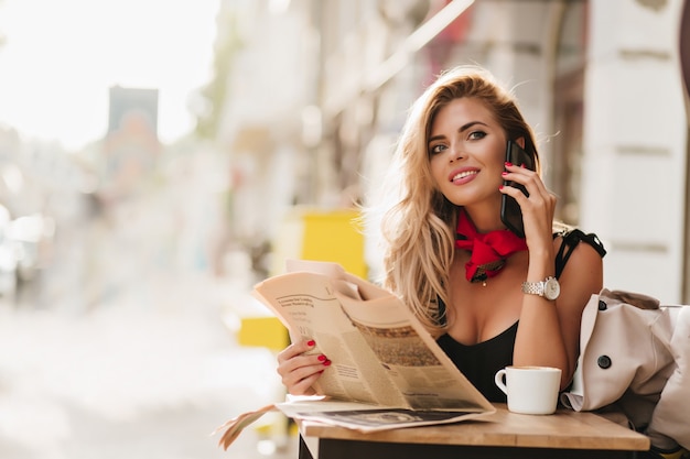 Восторженная кудрявая девушка смотрит с улыбкой во время разговора по телефону в летнем кафе