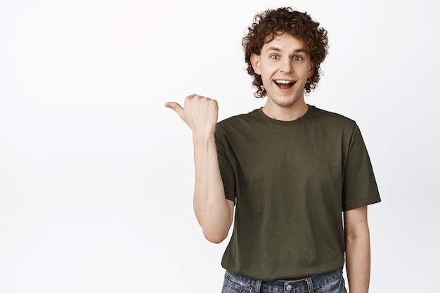 Enthusiastic curly guy pointing left smiling and looking happy while showing advertisement discount promo standing over white background