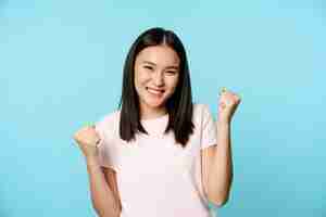 Free photo enthusiastic cute asian girl winning, rejoicing from great news, victory dance, smiling pleased, standing over blue background