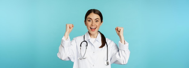 Enthusiastic doctor woman rejoicing winning and celebrating achieve goal standing in white coat agai