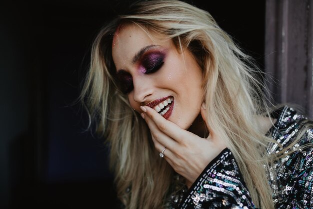 Восторженная европейская девушка с прямыми светлыми волосами смеется дома Модная фотосессия утонченной кавказской дамы с фиолетовым макияжем