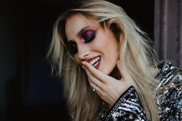 Восторженная европейская девушка с прямыми светлыми волосами смеется дома Модная фотосессия утонченной кавказской дамы с фиолетовым макияжем