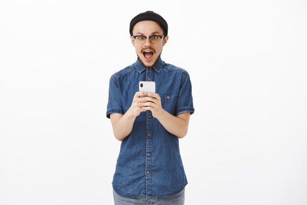 Enthusiastic and excited handsome creative male model with moustache in glasses black beanie dropping jaw from amazement and joy holding smartphone and receiving awesome unbelievable offer