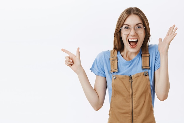Enthusiastic excited and thrilled emotive caucasian woman in glasses and brown overalls raising hand in surprise and joy pointing left smiling joyfully amazed and astonished over grey wall