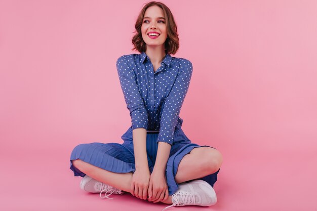 Восторженная женская модель с короткой стрижкой сидит на полу и смеется на розовой стене. Фотография в помещении: радостная европейка в синей блузке и белых кроссовках.