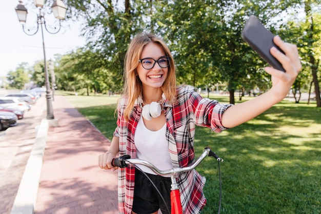 公園で自分撮りをする熱狂的な面白い女の子。自転車に乗って自分の写真を撮る素敵な金髪の女性モデル。