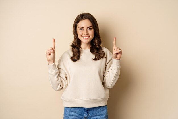 Enthusiastic girl pointing fingers up showing announcement promo text or logo upwards standing over ...