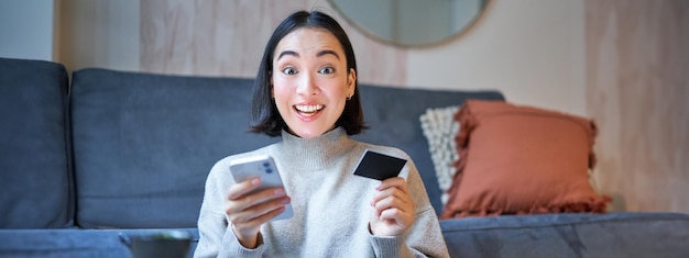 Free photo enthusiastic girl sits at home with smartphone and credit card pays for online order book tickets on