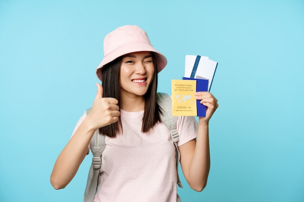 Enthusiastic girl traveller asian tourist shows thumbs up and international vaccine certificate heal...