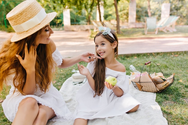 Восторженная девушка с длинными каштановыми волосами держит зеленое яблоко и разговаривает с мамой. Красивая женщина в элегантной шляпе, касаясь лица дочери пальцем, сидя на одеяле в парке.