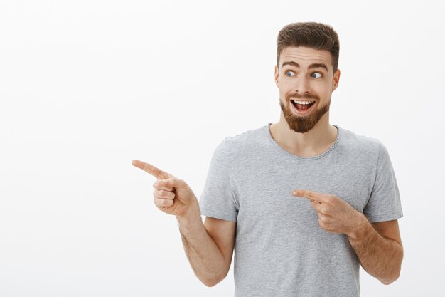 Enthusiastic guy feeling happy and amazed gazing at super cool copy space. Portrait of handsome delighted and charismatic man with beard and moustache looking and pointing left over gray wall