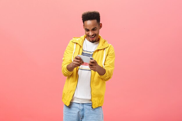 Enthusiastic guy having fun playing awesome smartphone game holding cellphone in both hands staring at device screen with passion and thrill spending free time in internet over pink background