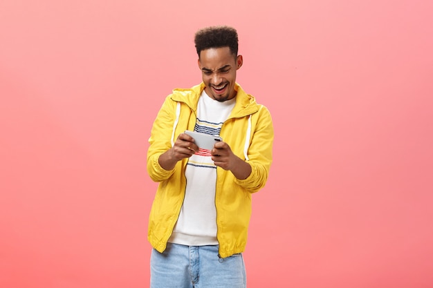 ピンクの背景の上でインターネットで自由な時間を過ごす情熱とスリルでデバイスの画面を見つめている両手で携帯電話を持って素晴らしいスマートフォンゲームを楽しんでいる熱狂的な男
