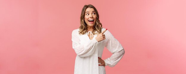 Enthusiastic happy attractive blond woman in white cute dress open mouth excited and cheerful looking pointing upper right corner as best prices discounts ever stand pink background