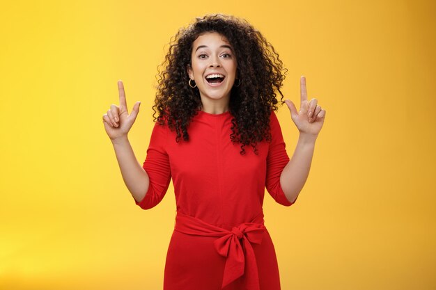 Enthusiastic happy curly-haired young european woman feeling happy present awesome copy space, raising hands pointing up and smiling with delight and admiration over yellow background.