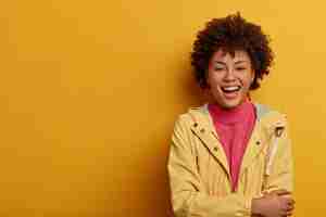 Free photo enthusiastic hilarious woman with afro hairstyle, laughs out loud, imagines funny situation, keeps hands crossed over chest, dressed casually, stands against yellow wall