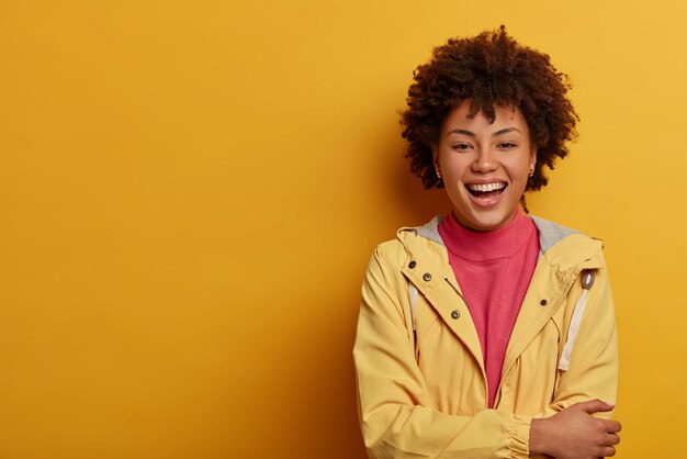 Enthusiastic hilarious woman with Afro hairstyle, laughs out loud, imagines funny situation, keeps hands crossed over chest, dressed casually, stands against yellow wall