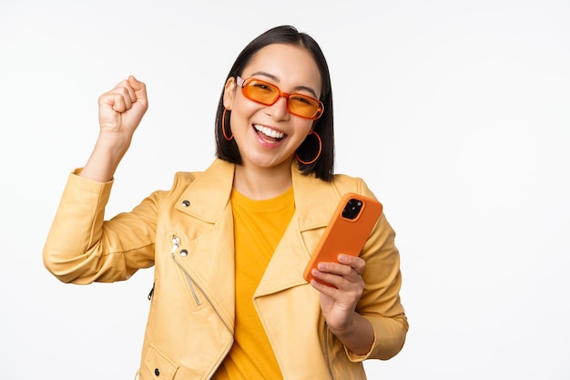Восторженная корейская девушка в солнцезащитных очках, держащая смартфон, празднует и танцует, смеясь, счастливая с мобильным телефоном, стоящим на белом фоне