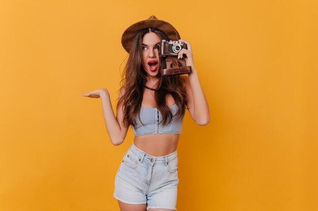 Enthusiastic long-haired brunette girl in hat holding camera and expressing positive emotions