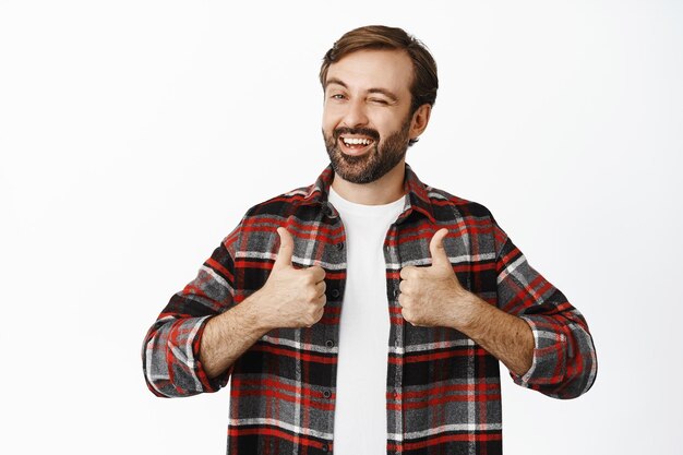 Enthusiastic male model winks and smiles shows thumbs up stands in checked shirt over white background