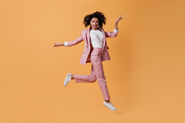 Enthusiastic mixed race girl jumping on yellow background Full length view of gorgeous young woman in pink suit