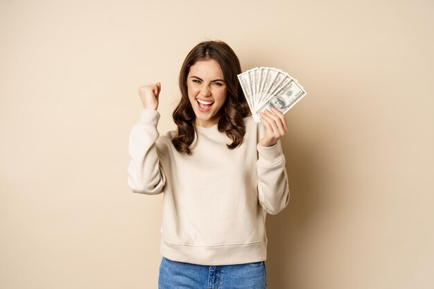 Enthusiastic modern woman winning money got cash celebrating and shouting of joy standing against be...