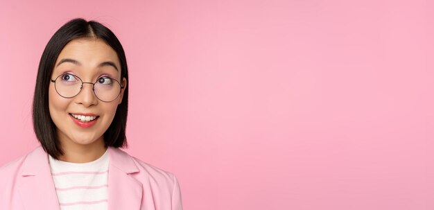 Enthusiastic saleswoman asian businesswoman in glasses looking intrigued at upper left corner logo company advertisement standing over pink background