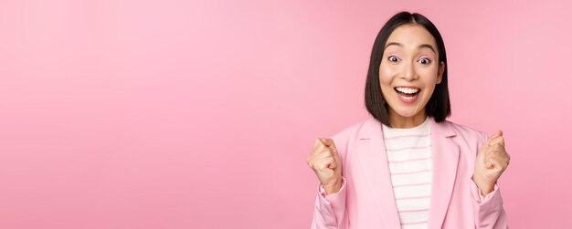 Enthusiastic saleswoman asian corporate woman say yes achieve goal and celebrating triumphing looking with rejoice and smiling standing over pink background